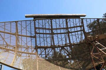 Large, vintage, parabolic mesh radar antenna or radio telescope dish set against a clear blue sky, likely outdoors in a park or museum.