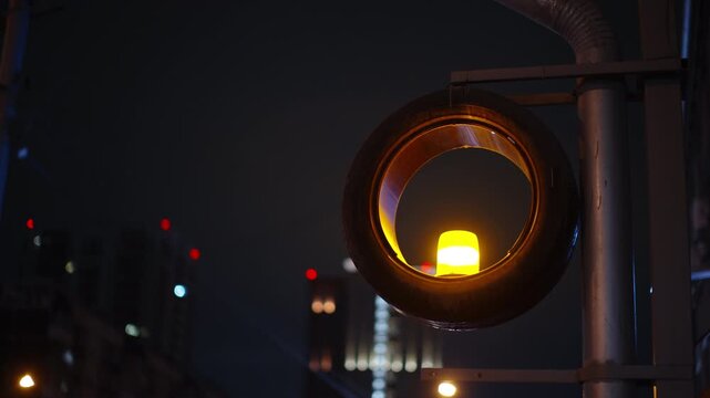 Urban night scene with a hanging tire encircling a yellow warning beacon on a metal pole, set against a distant city skyline with red-tipped towers and soft bokeh lights.