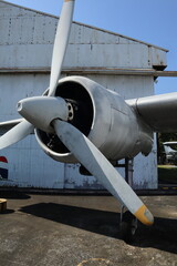 Close-up view of the engine and propeller of a large, silver vintage aircraft (likely a bomber or transport plane) parked in front of a corrugated metal hangar.