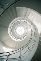 Fototapeta premium Architectural white spiral staircase with glass and steel railing, viewed from above, centering on a bright circular skylight.