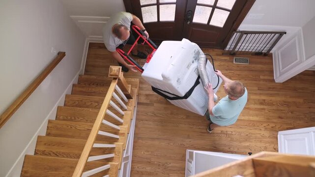 caucasian moving crew members coordinating two-person lift of washing machine at base of stairs, synchronized movements, red dolly and protective straps visible, emphasis on precision and care