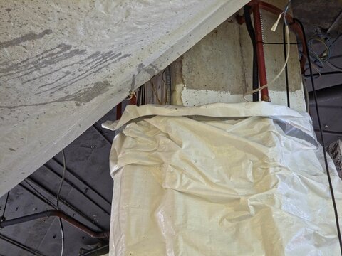 Low-angle, abstract view of a construction site featuring a streaked concrete ceiling, a column wrapped in crinkled white plastic, and exposed wiring and pipes overhead. - Powered by Adobe