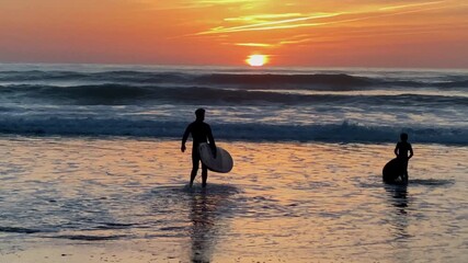 Two surfers entering the water under a dramatic orange sunset sky over the wide sandy beach of Costa Ballena with calm gentle waves - Powered by Adobe