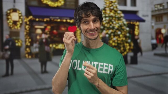 Man holding red heart card in green volunteer shirt on street with blurred holiday lights; joy compassion.
