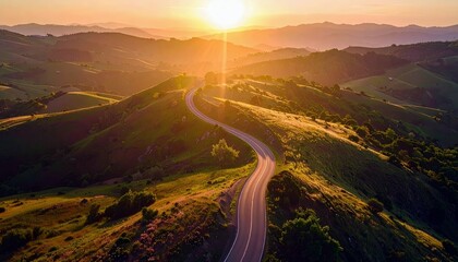 Aerial view of a winding road cutting through lush green hills, bathed in the warm glow of a setting sun, creating a scenic landscape.