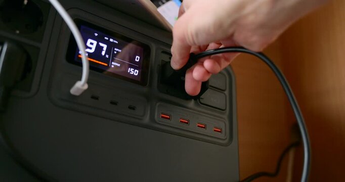 A hand is shown plugging a black power cord into a portable power station with a digital display.