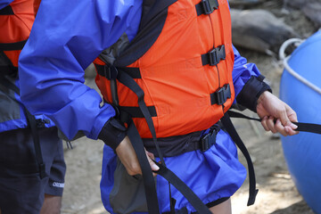 Person in blue jacket and bright orange life vest preparing for water adventure. Focused close up of hands adjusting black safety strap for fun river rafting activity