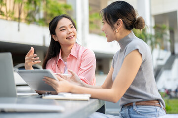 Fototapeta premium Asian woman student talking to friend holding tablet working on group project at table in university