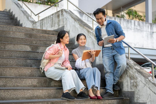 Asian man student standing showing tablet to woman friends sitting on building stairs in university.