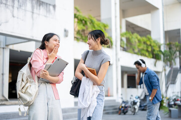 Fototapeta premium Asian woman student holding laptop laughing with friend holding book standing walking in university.