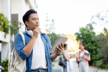Fototapeta premium Asian man student carrying a bag holding pen looking away from tablet while standing in university.