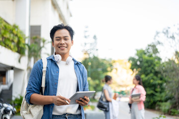 Smiling asian man student carrying a bag holding pen and tablet while standing in the university.