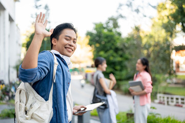 Happy asian man student holding tablet waving a hand while walking to woman friends in university