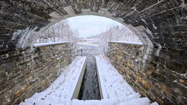 A spillway passes under a stone arch bridge into a creek that flows toward the Ottawa River, seen on a snowy winter day - Powered by Adobe