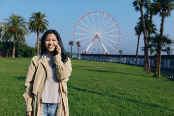 Woman in a beige coat smiles while talking on her phone in a sunny park, green grass and a distant...