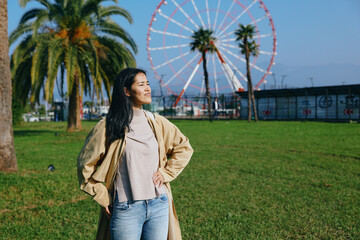 Woman in a beige cape and casual sweater stands in a sunny park with a ferris wheel in the...