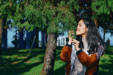 Nature outdoors scene with a smiling woman smelling greenery under a tree in a sunlit park, wearing...