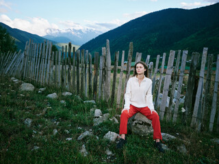 Fototapeta premium Woman sits beside a weathered wooden fence in a mountainous landscape, wearing red pants and a light sweater, with green grass, rocks, and distant snowcapped peaks.
