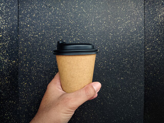 Hand holding a cup of coffee. Hand holding blank brown disposable paper coffee cup mockup with black lid and copy space for branding design.