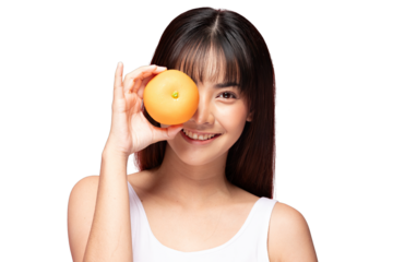 Asian Young woman smiling while holding orange fruit over one eye, fresh healthy skin, casual white tank top, bright studio portrait with playful expression