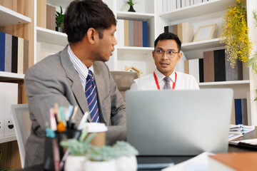Two men are sitting at a desk, one of them is holding a piece of paper. They are having a conversation, and it seems like they are discussing something important. The atmosphere is tense
