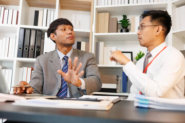 Two men are sitting at a desk, one of them is holding a piece of paper. They are having a conversation, and it seems like they are discussing something important. The atmosphere is tense
