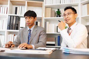 Two men are sitting at a desk, one of them is holding a piece of paper. They are having a conversation, and it seems like they are discussing something important. The atmosphere is tense
