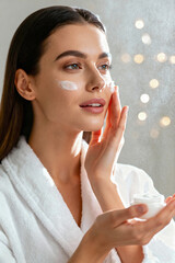 Beautiful young woman in a white bathrobe applying moisturizing cream to her cheek in an indoor setting with sparkling bokeh lights in the background