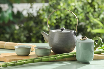 Board with teapot, cups of tea and bamboo stems on wooden table outdoors