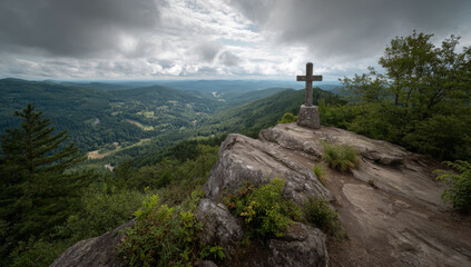 Cross atop a rocky peak overlooking a lush valley under a cloudy sky.