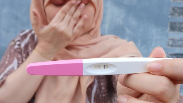 A young Asian muslim woman covers her face with her hand and cries, in her other hand there is a negative pregnancy test. A woman's desire to have children. 