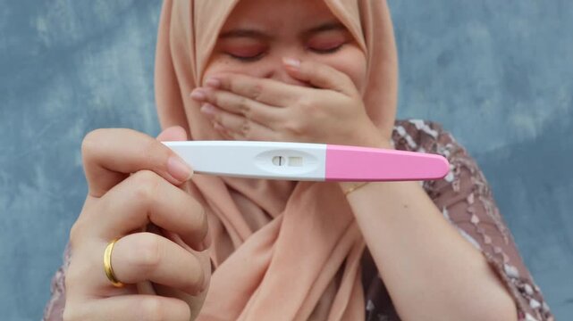 A young Asian muslim woman covers her face with her hand and cries, in her other hand there is a negative pregnancy test. A woman's desire to have children. 