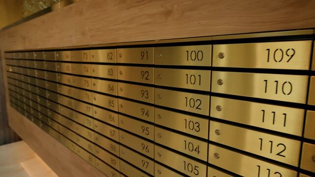 Warm-lit row of brass numbered mailboxes built into a wooden wall in a residential lobby, with horizontal compartments and sequential numbering for postal delivery and storage