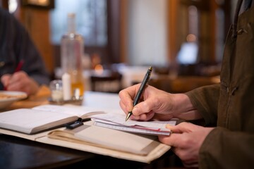 Man Writing in Notebook at Table with Whiskey Bottle