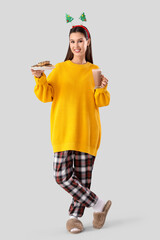 Beautiful young woman in pajamas with glass cup of hot cocoa and sweet cookies on grey background. Christmas celebration