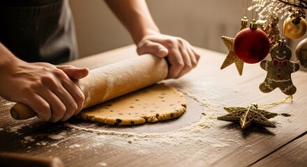 Hands rolling out cookie dough on a wooden surface surrounded by Christmas decorations.