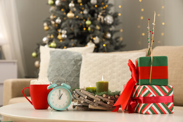 Alarm clock with cup of cacao, candles and gift boxes on coffee table near Christmas tree in living room