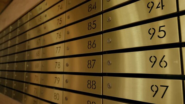 Rows of brass numbered mailboxes and locker compartments lining a building corridor, in a close-up perspective, showing engraved numbers on slots arranged in a clean, uniform grid.