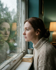 Pensive woman gazing at rain-streaked window in dimly lit room.