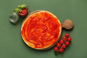 Board with raw dough for pizza and jar of tomato sauce on green background