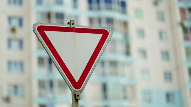 Close-up view of a triangular yield traffic sign with a bold red border and white center, set before a blurred facade of residential apartments in an urban street