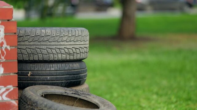 Stack of worn automotive tires leaning against a brick wall on a grassy lawn, with a blurred park background, highlighting rubber tread texture and themes of recycling and reuse.