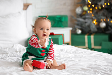 Naklejka premium Cute little baby in elf costume with gift box sitting on bed in bedroom. Christmas celebration