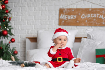 Cute little baby in Santa costume with mini Christmas trees sitting on bed in bedroom