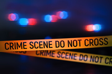 Closeup view of yellow crime scene tape with police car lights flashing in the background at night. Focus on the tape with bokeh, blur and lens glow creating dramatic investigation mood