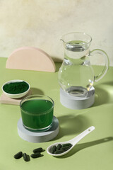 Glass of spirulina drink, jug of water and pills on green table, closeup