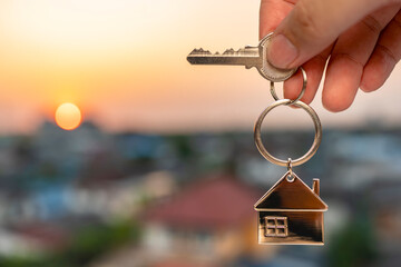 Hand holding a house keychain with a key against sunset sky, symbolizing new home ownership, real...