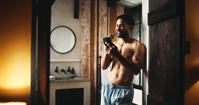 Phone, brushing teeth and man in home for dental wellness, hygiene and morning news. Mobile, cleaning mouth and shirtless person by bathroom doorway for oral care, health and reading message online