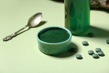 Bowl with spirulina powder, pills and spoon on green background