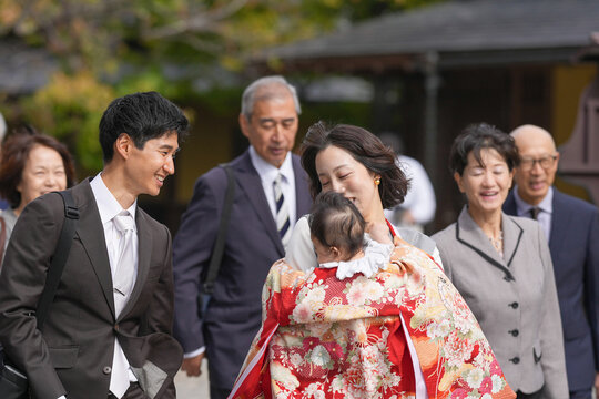 A man in a suit is smiling at a woman holding a baby. The baby is wearing a red and white outfit. The scene appears to be a family gathering or a special event